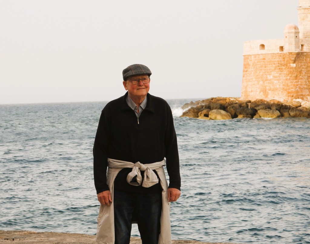 Ein älterer Mann steht am Meer, trägt einen grauen Hut und einen Pullover, mit einem Felsen im Hintergrund.