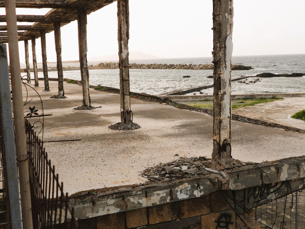 Verfallenes Gebäude mit sichtbaren Betonpfeilern und Meeresblick im Hintergrund.