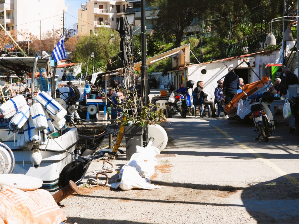 Landschaft eines Hafens mit Fischerbooten und Männern, die an einem Tisch sitzen. Im Hintergrund sind Wohngebäude und eine griechische Flagge sichtbar.