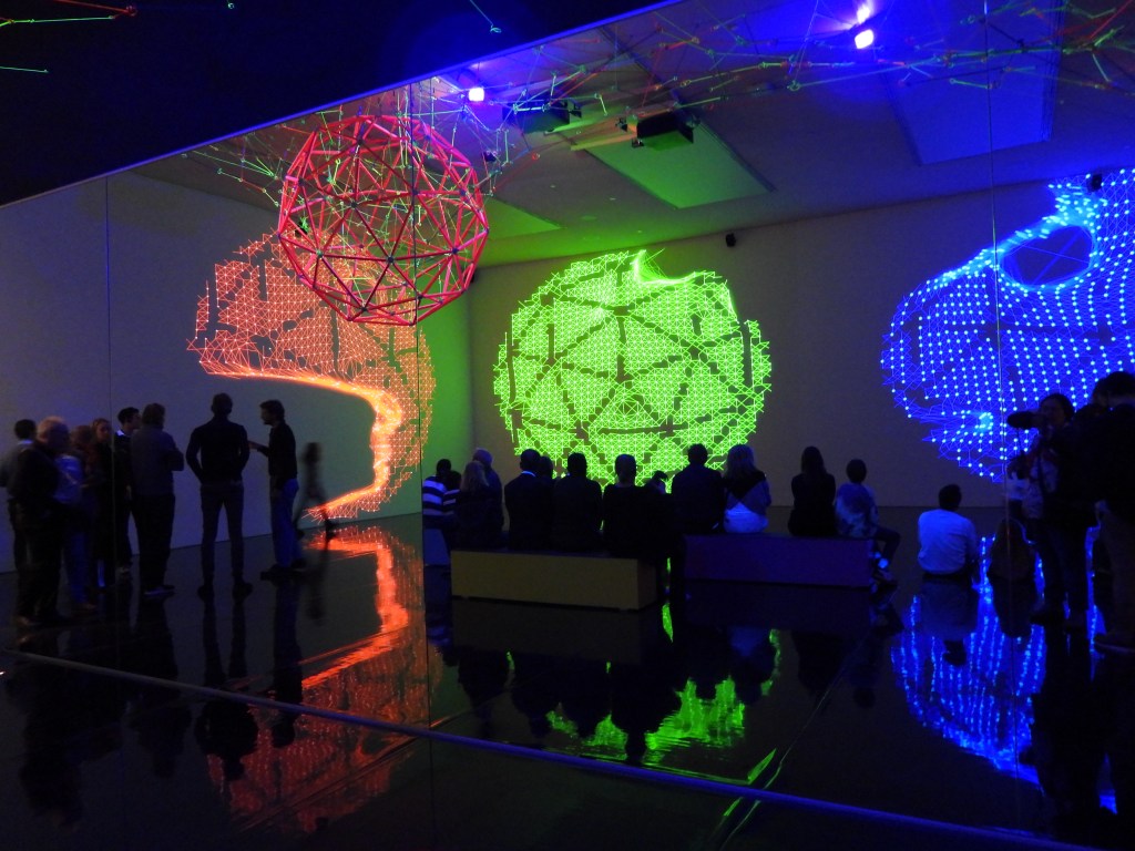 Eine Kunstausstellung mit bunten Lichtinstallationen, die geometrische Muster an die Wände projizieren. Gruppen von Menschen beobachten die visuelle Darstellung und reflektieren auf dem glänzenden Boden.