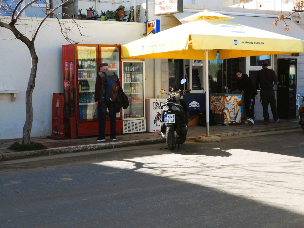 Eine Straße mit einem Motorrad und einem Kiosk unter einem gelben Sonnenschirm. Im Hintergrund steht ein Getränkeautomat und eine Person kauft etwas. Es sind auch andere Personen im Kiosk beschäftigt.