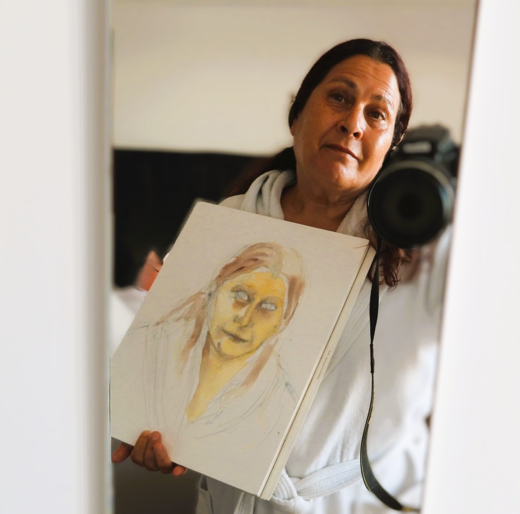 Eine Person in einem Bademantel hält eine Zeichnung mit einem Porträt einer Frau und schaut in einen Spiegel, während sie eine Kamera hält.