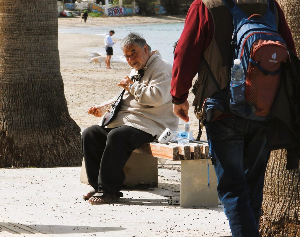 Ein älterer Mann sitzt auf einer Parkbank und spielt ein Musikinstrument, während ein Passant im Hintergrund vorbeigeht.