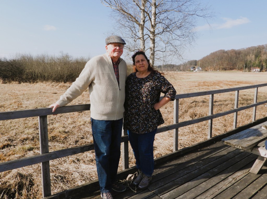 Ein älterer Mann und eine Frau stehen zusammen auf einer Holzplattform im Freien, umgeben von einem weiten, trockenen Feld und Bäumen im Hintergrund.
