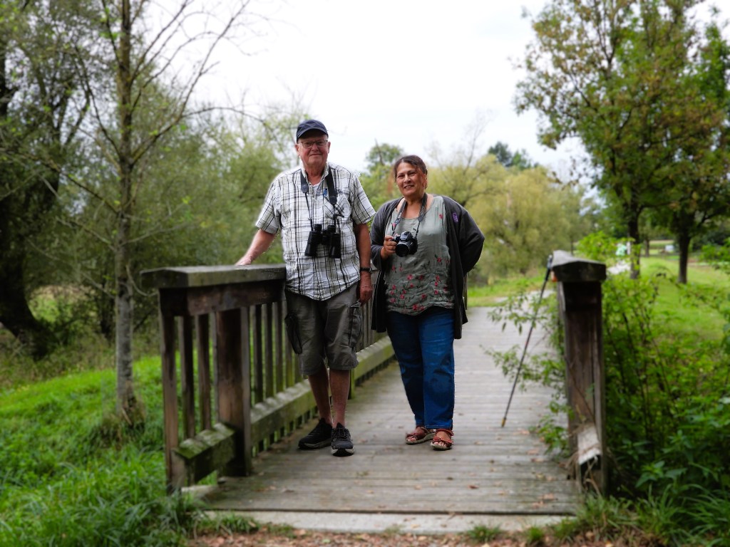 Zwei Menschen stehen auf einer Holzbrücke in einer grünen Umgebung und halten Kameras.