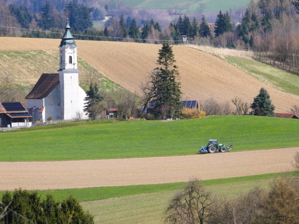 Landschaft mit einer Kirche und einem Traktor auf einem Feld, umgeben von Wäldern und hügeliger Wiese.