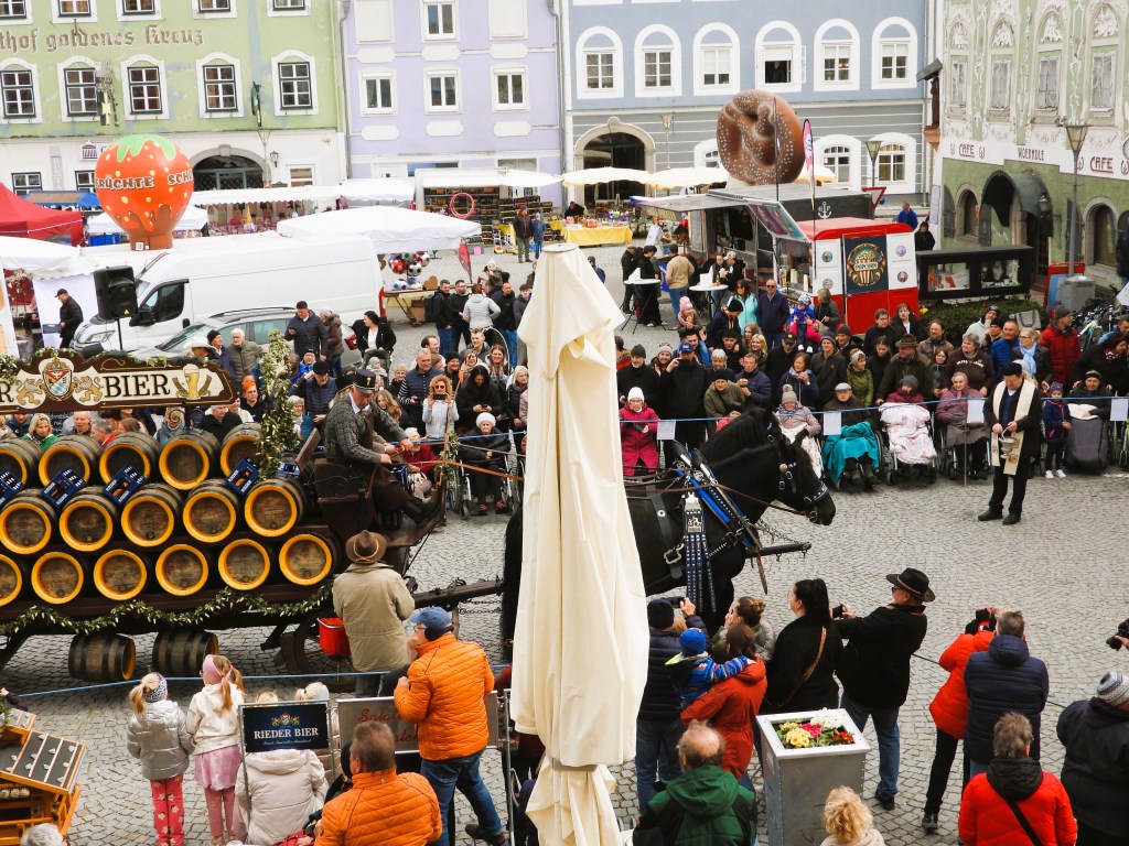 Menschenmenge auf einem Festivalplatz mit einem Pferd und einem Wagen, der mit Bierfässern beladen ist. Zahlreiche Zuschauer beobachten das Geschehen, während Stände im Hintergrund eine festive Atmosphäre schaffen.