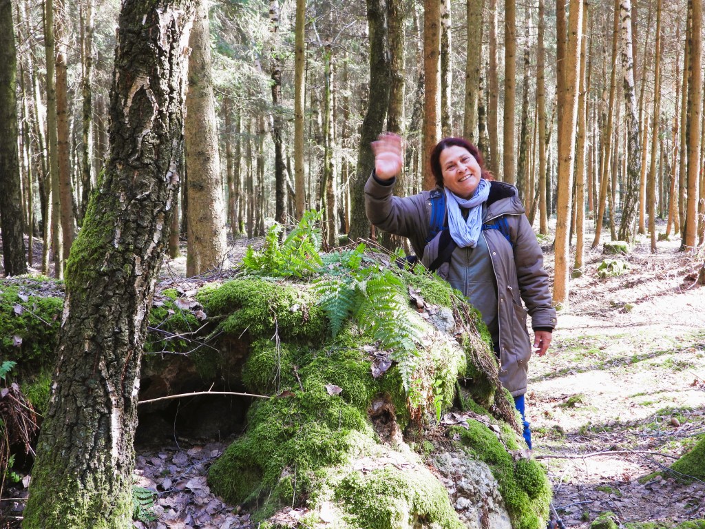 Eine Frau winkt fröhlich in einem Wald mit hohen Bäumen und moosbedecktem Boden.
