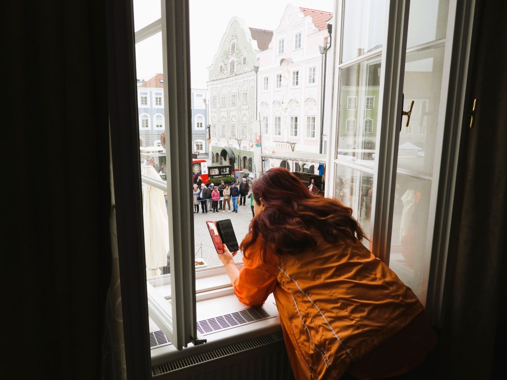 Eine Person mit langen, braunen Haaren in einer orangefarbenen Jacke lehnt sich aus einem offenen Fenster und schaut auf einen Platz mit Menschen und historischen Gebäuden.