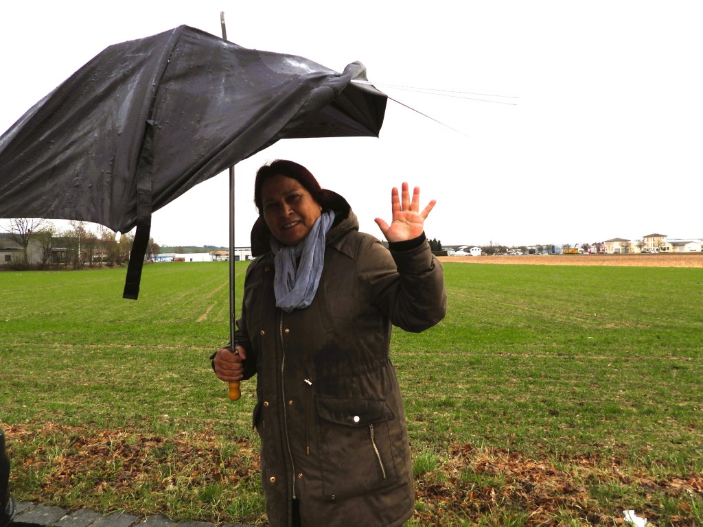 Eine Frau in einem braunen Mantel hält einen umgedrehten Regenschirm in der Hand und winkt lächelnd. Im Hintergrund ist ein grünes Feld und ein grauer Himmel zu sehen.