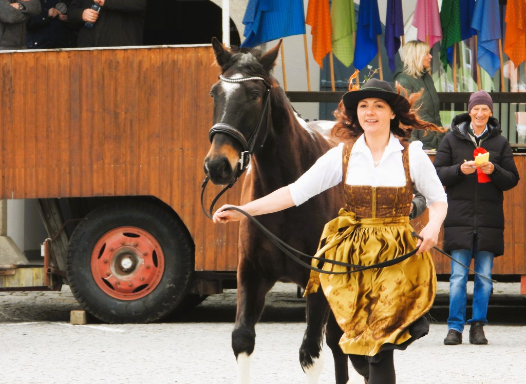 Eine Frau in traditioneller Kleidung führt ein Pferd, während Zuschauer im Hintergrund stehen.