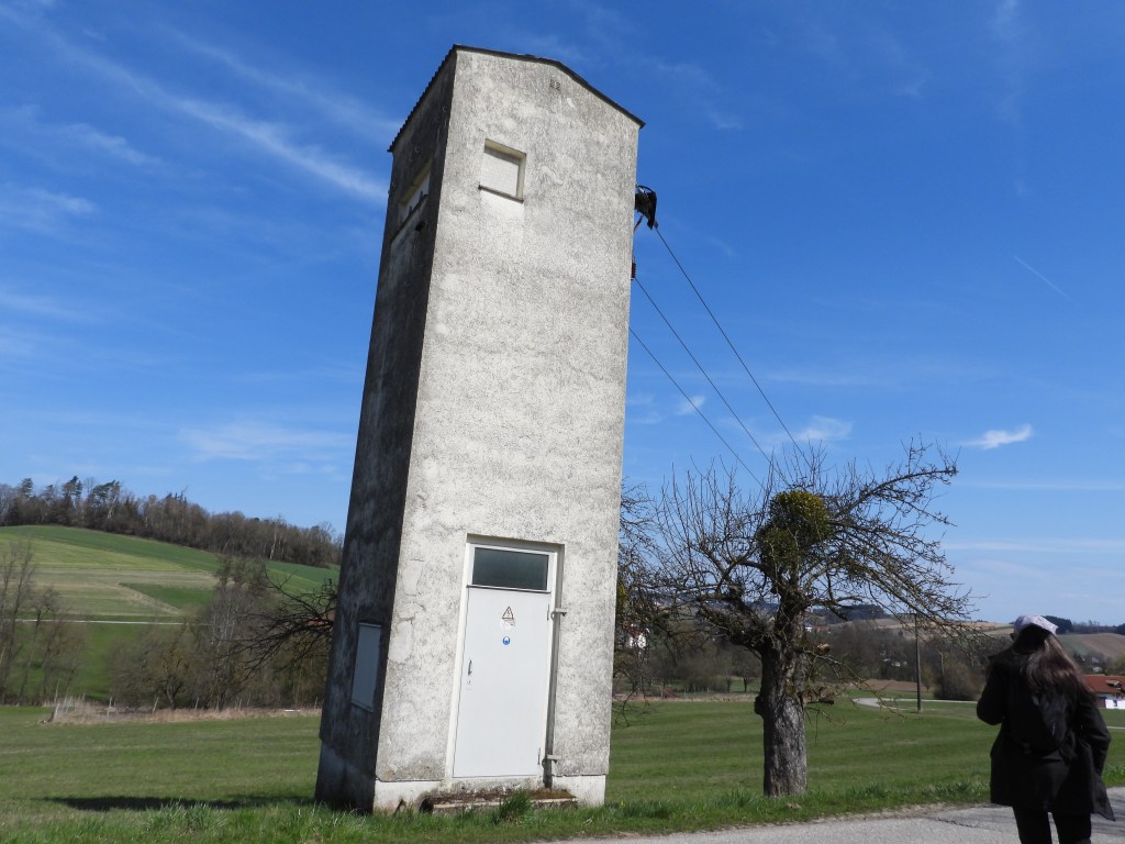 Ein schmaler, hoher, grauer Turm steht auf einer Wiese, umgeben von verschiedenen Grüns und blauen Himmel. Im Vordergrund sieht man eine Baum und zwei Personen, die sich der Szenerie nähern.