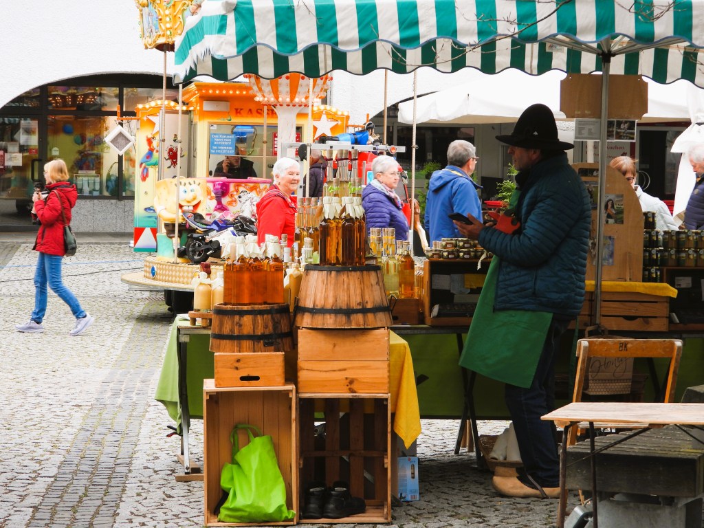 Marktstand mit Flaschen und Fässern, während Passanten im Hintergrund schlendern.