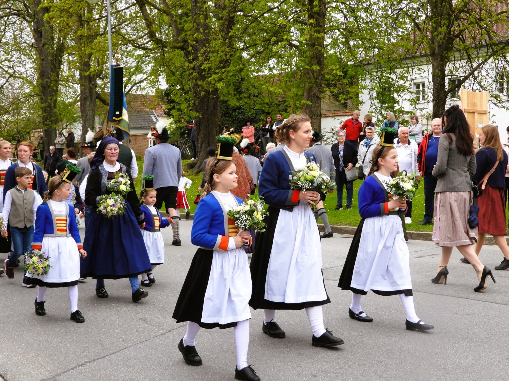Eine Gruppe von Personen in traditioneller Kleidung, die an einem Festumzug teilnimmt. Im Vordergrund gehen Frauen und Mädchen mit Blumensträußen, während Zuschauer im Hintergrund stehen.