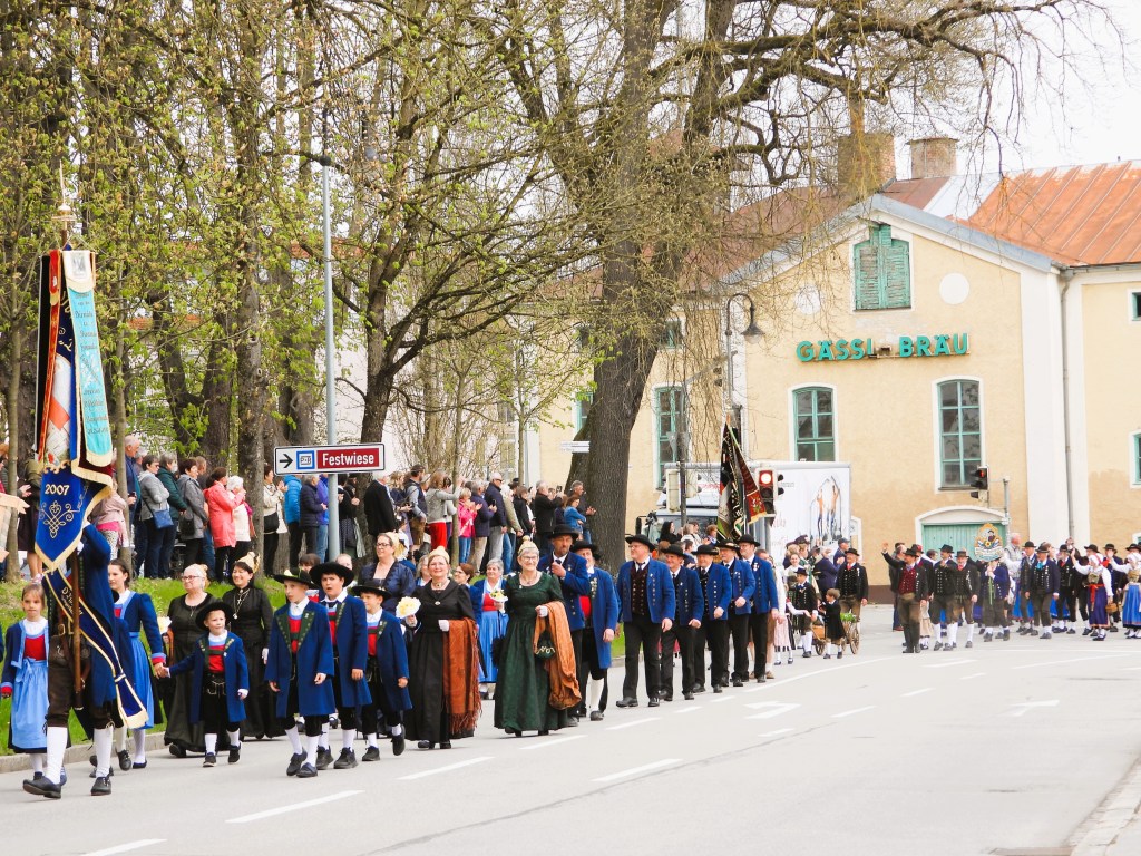 Eine Festparade mit Menschen in traditionellen Trachten, die auf einer Straße marschieren, während Zuschauer auf den Bürgersteigen stehen. Im Hintergrund ist ein Gebäude mit der Aufschrift 'GASSI-BRÄU' zu sehen.