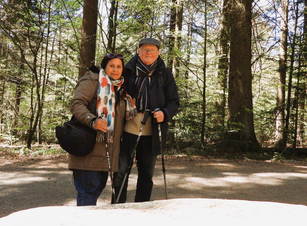 Ein Paar steht in einem Wald, umgeben von Bäumen, während sie Wanderstöcke halten. Die Frau trägt einen langen Mantel und einen bunten Schal, der Mann hat einen Hut und einen Schal an.