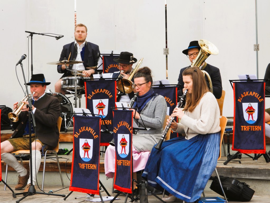 Eine Blaskapelle spielt auf einer Bühne. Musiker mit verschiedenen Instrumenten, darunter Trompete, Klarinette und Schlagzeug, tragen traditionelle Kleidung. Im Hintergrund sind Banner mit dem Schriftzug 'Blaskapelle Triftern' zu sehen.