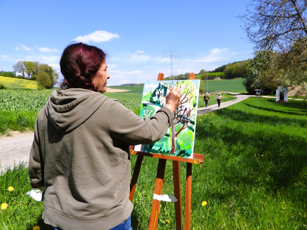 Frau malt an einer Leinwand im Freien, umgeben von einer grünen Landschaft mit blauen Himmel und Wolken.