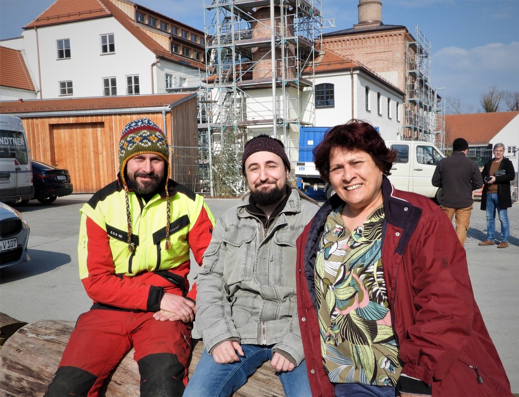Drei Personen sitzen auf einem Baumstamm vor einem Gebäude im Hintergrund, das im Umbau ist. Die erste Person trägt eine gelbe Arbeitsjacke und einen bunten Hut, die zweite Person hat einen dunklen Pulli und einen Bart, die dritte Person trägt eine rot-braune Jacke und ein gemustertes Oberteil.
