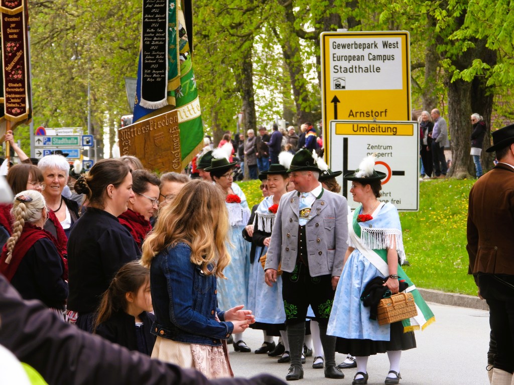Eine Gruppe von Menschen, die traditionelle Trachten tragen, steht an einer Straße mit einem Verkehrsschild. Sie interagieren freundlich, während sie an einer Veranstaltung teilnehmen.