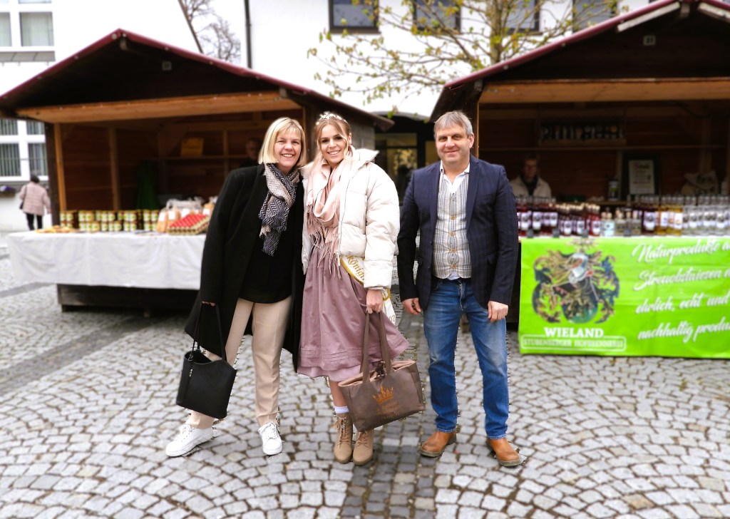 Drei Personen stehen lächelnd vor einem Marktstand mit Produkten im Freien.