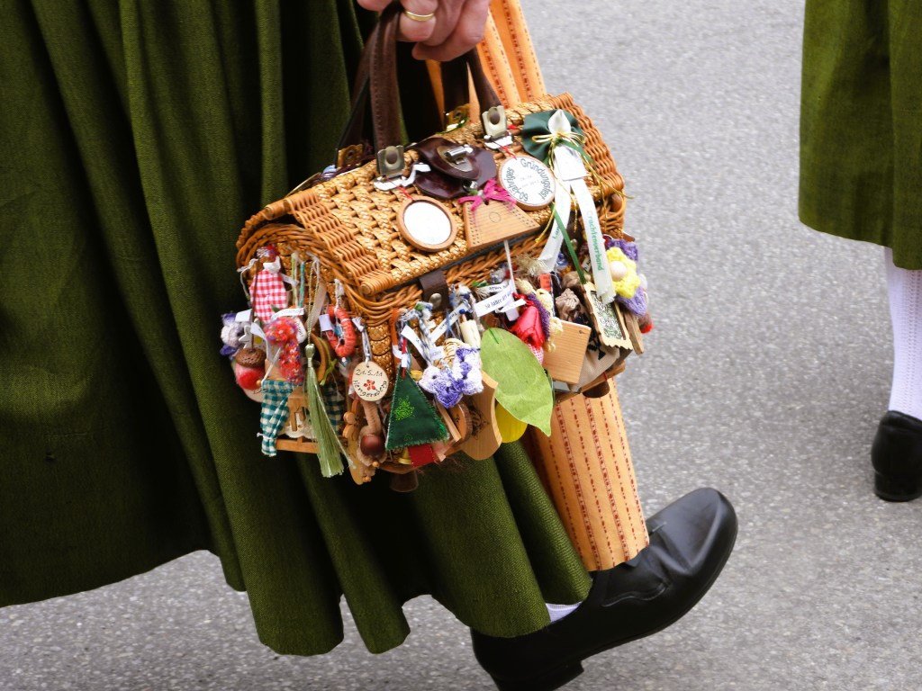 Nahaufnahme einer handgefertigten Korbtasche, dekoriert mit bunten Anhängern und kleinen Accessoires, getragen von einer Person in traditioneller Tracht.