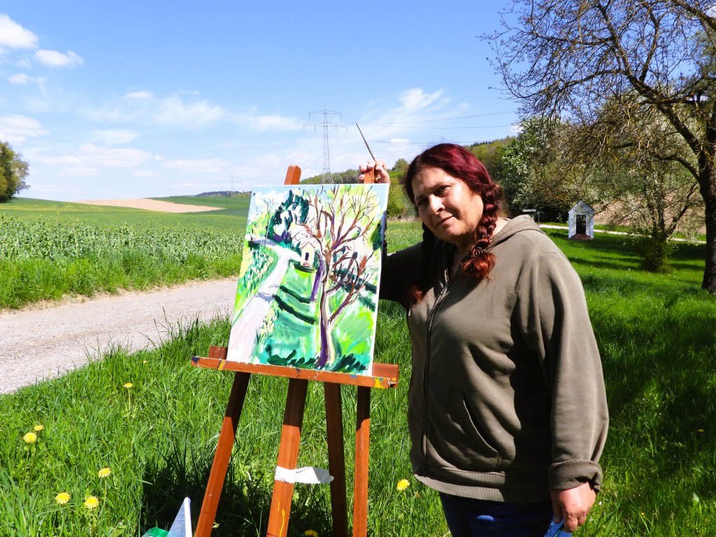 Eine Frau steht im Freien neben einem Staffelei mit einem Gemälde, umgeben von grüner Wiese und Landschaft.