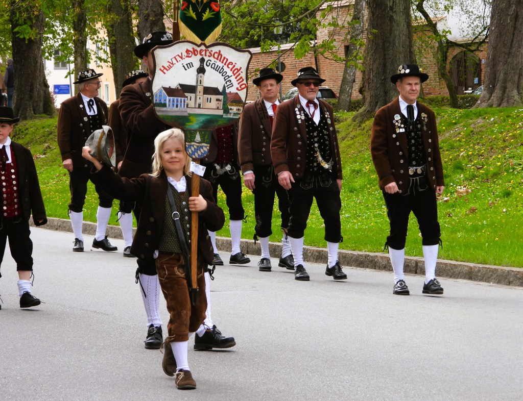 Eine Gruppe von Menschen in traditionellen österreichischen Trachten, die an einem Umzug teilnehmen. Ein Junge vorne hält ein großes Schild, während die anderen Teilnehmer in gleichfarbiger Kleidung folgen.