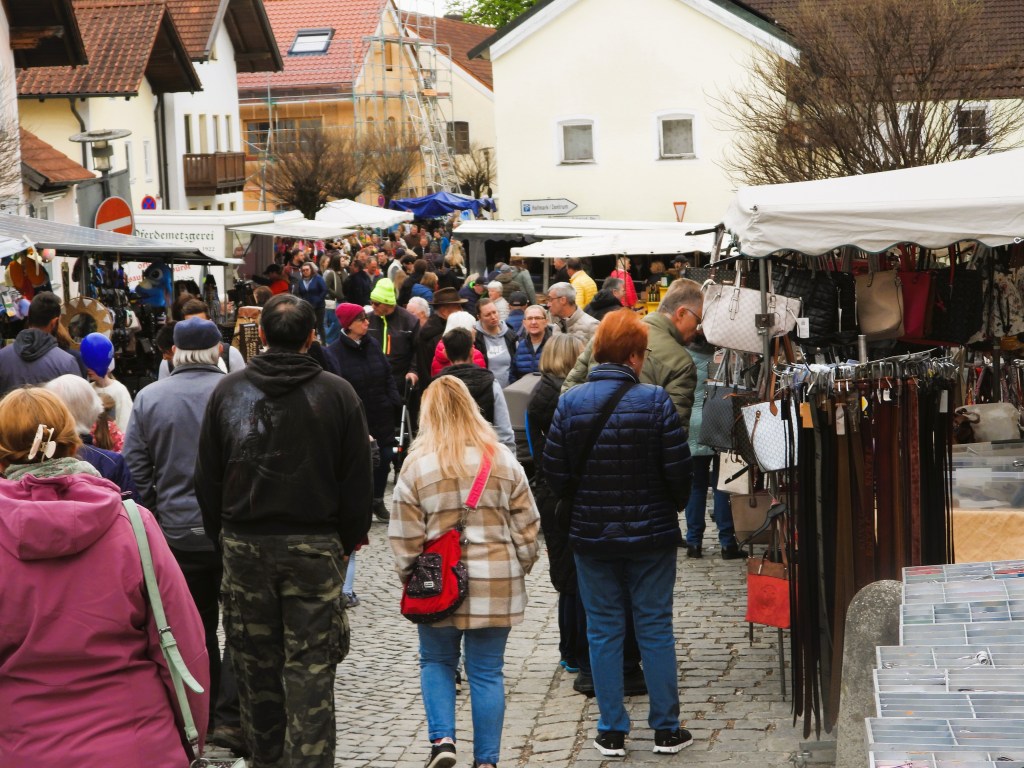 Belebter Markt mit vielen Menschen, die zwischen Ständen mit Taschen und anderen Waren schlendern. Im Hintergrund sind Gebäude und Bäume sichtbar.