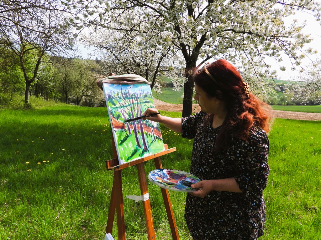 Eine Künstlerin malt an einem easel im Freien, umgeben von blühenden Bäumen und grünem Gras.