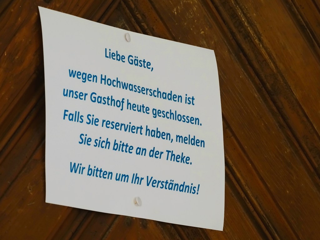 Ein Hinweis auf einem Blatt Papier, das an einer Wand hängt. Es informiert Gäste, dass der Gasthof aufgrund von Hochwasserschäden geschlossen ist und fordert Reservierungen zur Meldung an der Theke auf. Der Text endet mit einer Bitte um Verständnis.