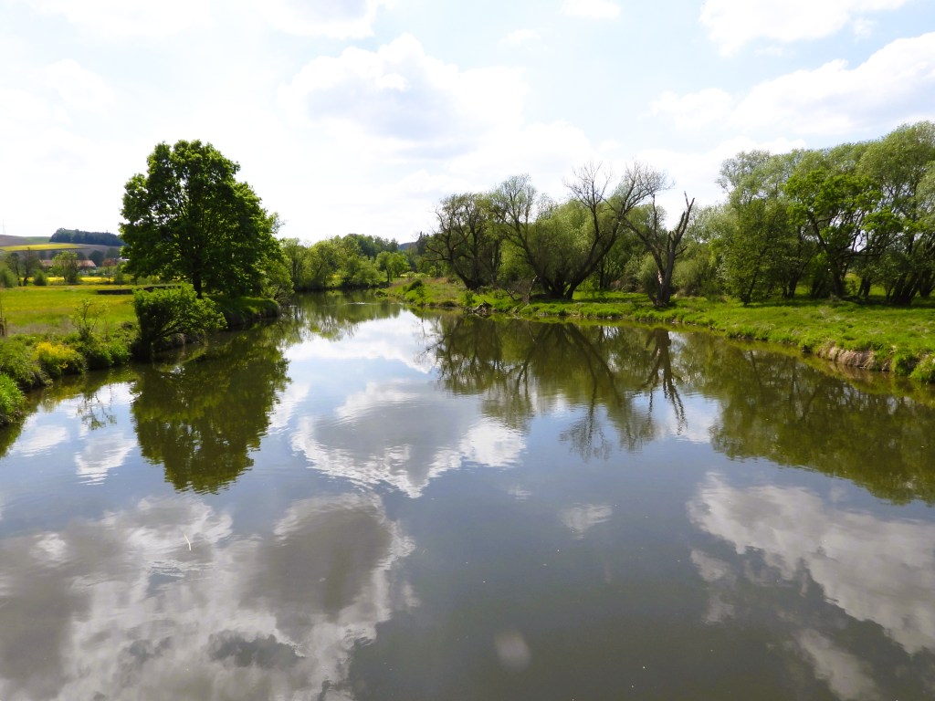 Ein ruhiger Fluss, der von üppigem Grün und Bäumen gesäumt ist, spiegelt den Himmel und die Wolken wider.