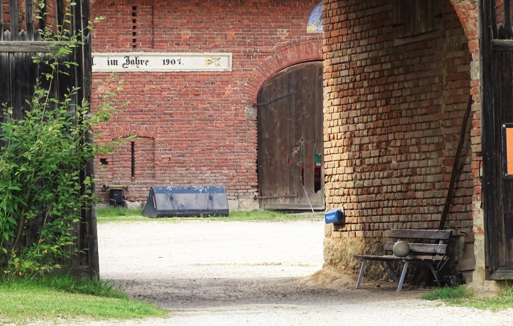 Blick durch einen Torbogen aus Ziegeln mit einer Bank im Vordergrund und einem historischen Gebäude im Hintergrund, das das Jahr 1907 zeigt.