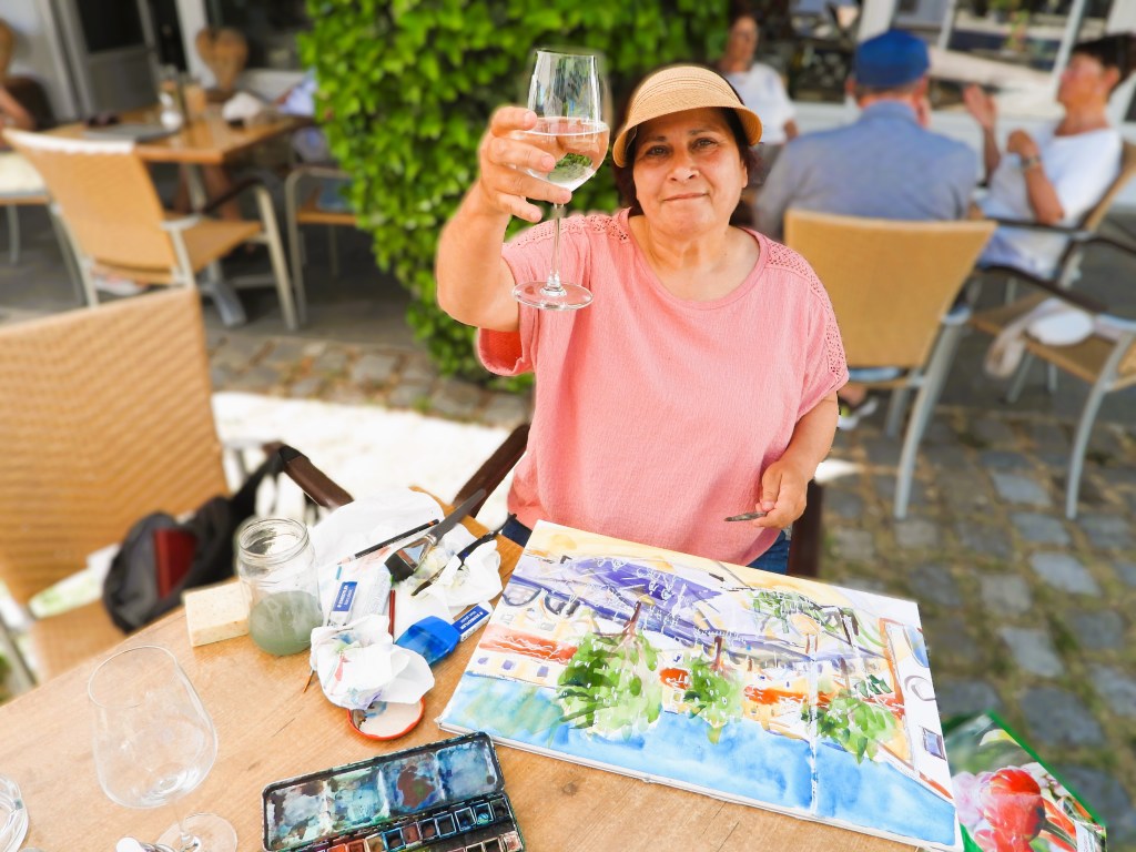 Frau mit Strohhut, die ein Glas Wasser hebt, während sie an einem Tisch im Freien sitzt und ein Aquarellbild malt. Künstlerin aus Bad Birnbach