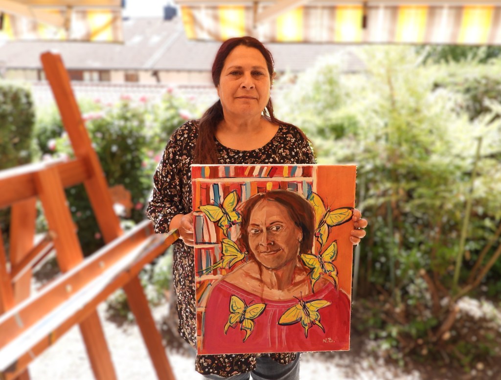 Eine Frau steht im Freien und hält ein buntes Gemälde mit Schmetterlingen und einem Porträt von sich selbst in den Händen.