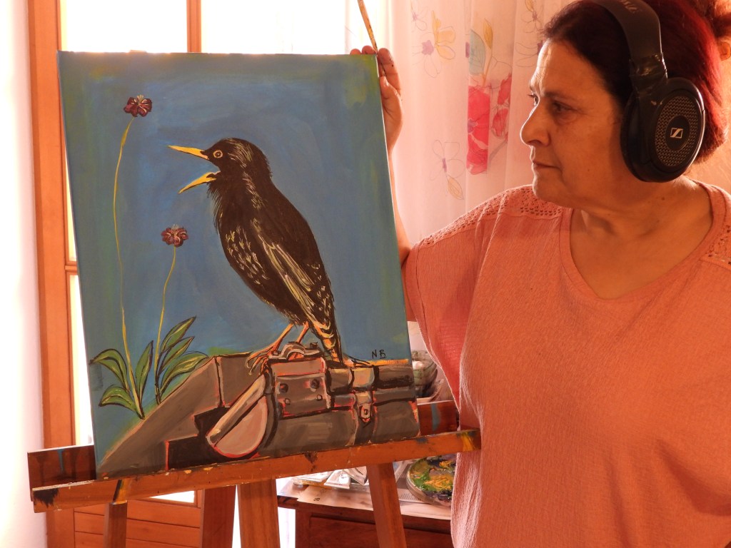 Eine Frau in Kopfhörern hält ein Gemälde eines singenden Vogels vor einem blauen Hintergrund, während sie an einer Staffelei steht.