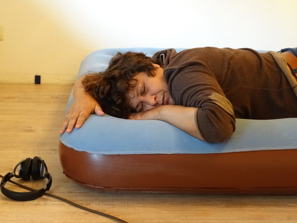 Eine Person schläft auf einer Luftmatratze, mit dem Kopf auf den Händen liegend. Neben der Matratze liegen Kopfhörer.
