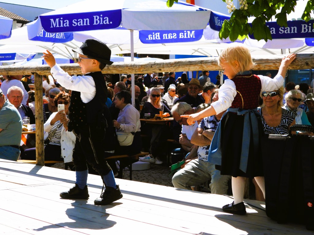 Zwei Kinder in traditioneller Tracht tanzen auf einer Bühne, während eine Menge von Zuschauern mit Bier und Essen auf Bänken sitzt. Sonnenschein und Schirme im Hintergrund.