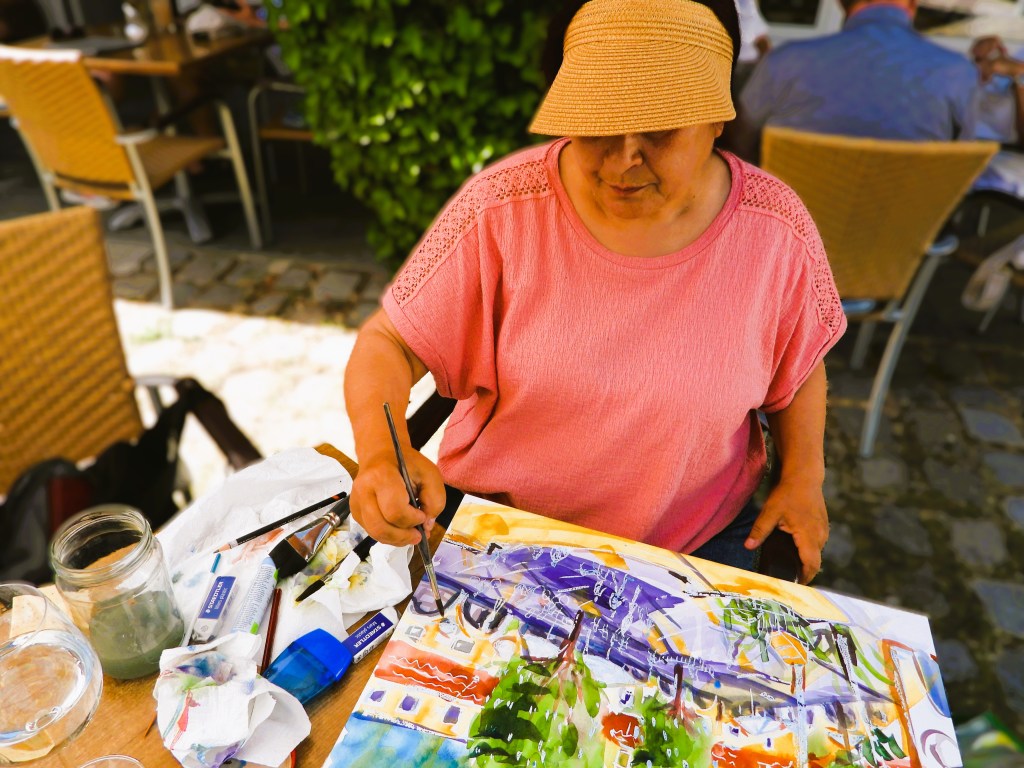 Eine Frau in einem pinken Shirt malt leidenschaftlich an einem Tisch im Freien, umgeben von Malutensilien und einer mediterranen Umgebung.