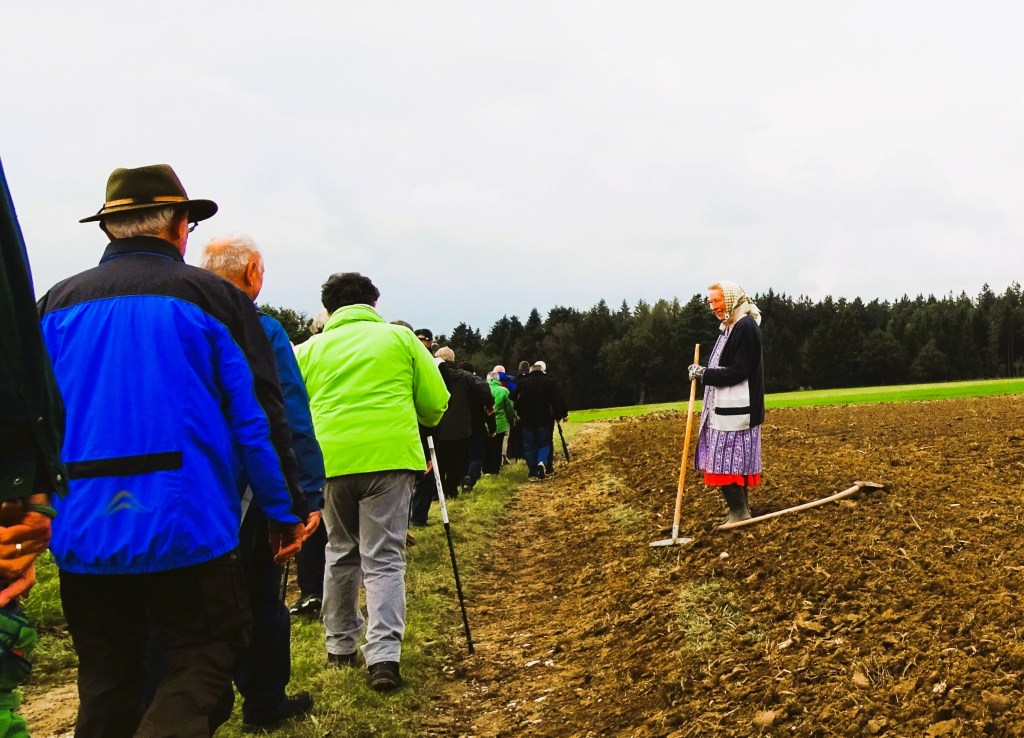 Eine Gruppe von Menschen geht in einer Reihe auf einem Feld, während eine ältere Frau mit einem Pflug steht.
