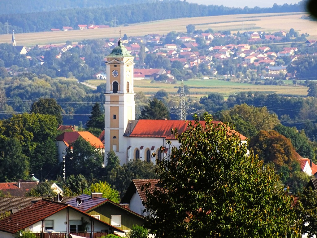 Blick auf eine Kirche mit Uhrturm in einer ländlichen Umgebung, umgeben von einem Dorf und Feldern im Hintergrund.