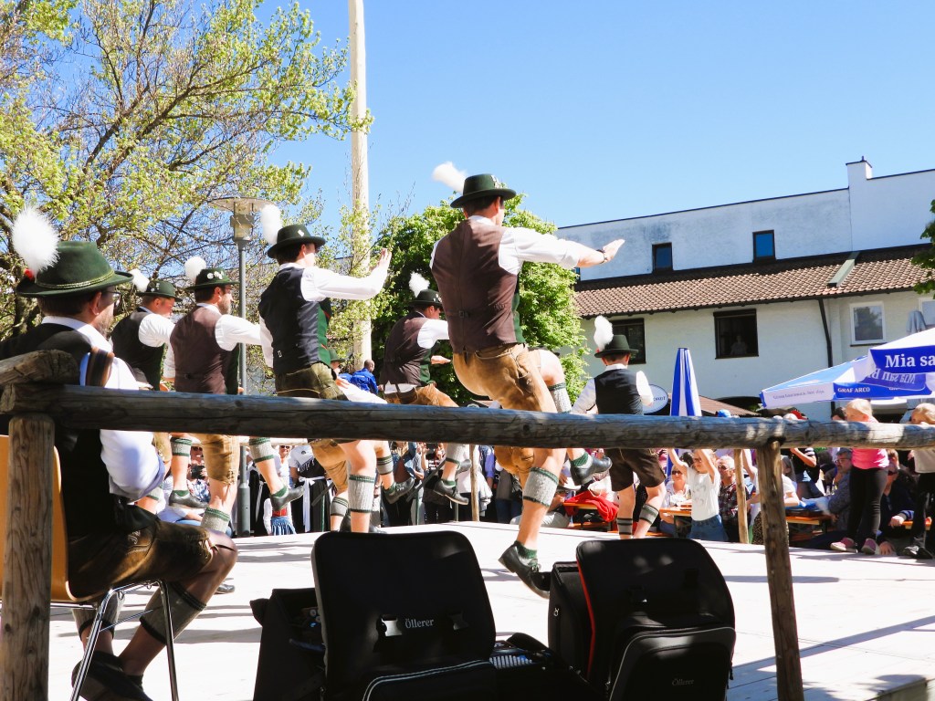 Tanzende Männer in traditioneller bayerischer Tracht bei einem Fest im Freien, mit einem Publikum im Hintergrund unter blauem Himmel.