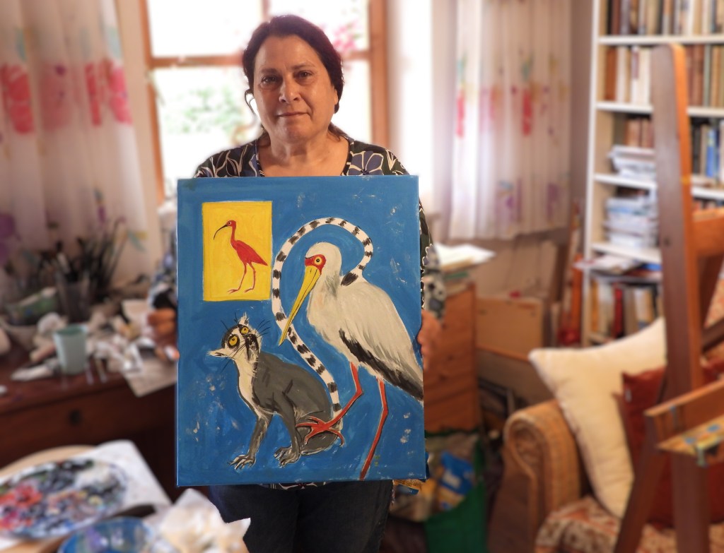 Eine Frau hält ein Gemälde mit einem Vogel, einem Storch und einer Comicfigur in einem Innenraum mit Kunstmaterialien im Hintergrund.