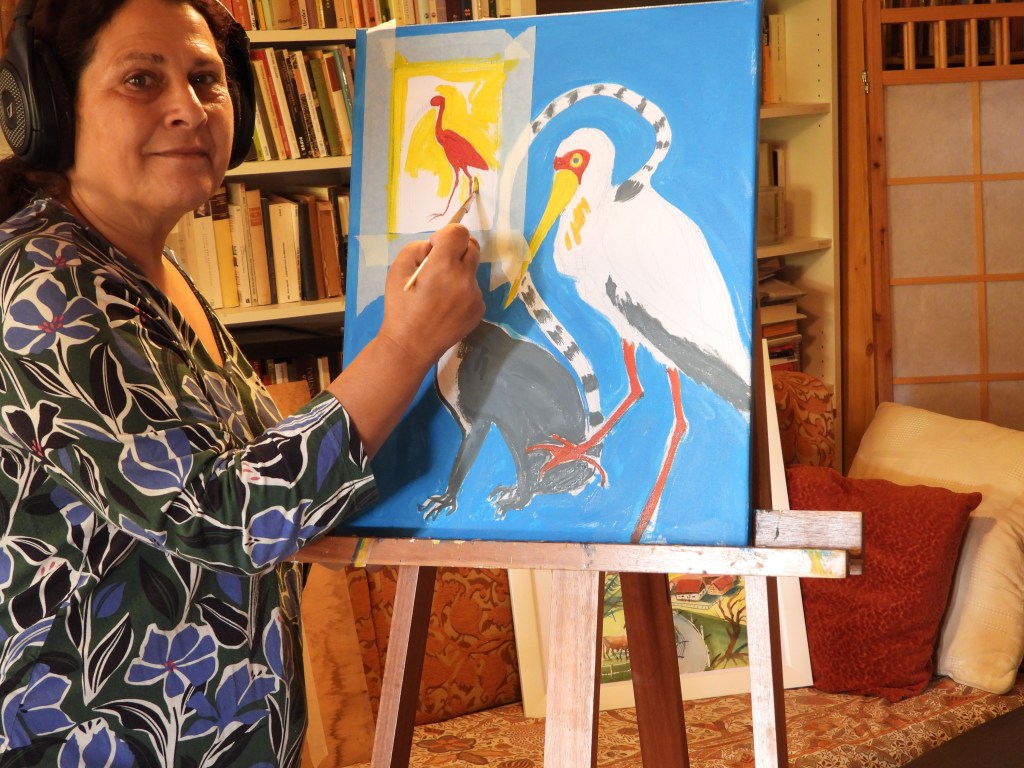 Eine Frau mit Kopfhörern malt auf einer Leinwand mit bunten Vögeln in einem gemütlichen Raum voller Bücher und Kissen.