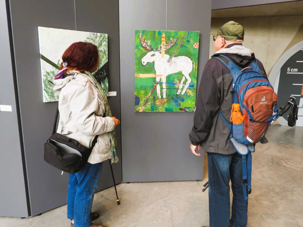 Zwei Personen betrachten Kunstwerke in einer Galerie, darunter ein farbenfrohes Gemälde eines Elches auf grünen Hintergrund.