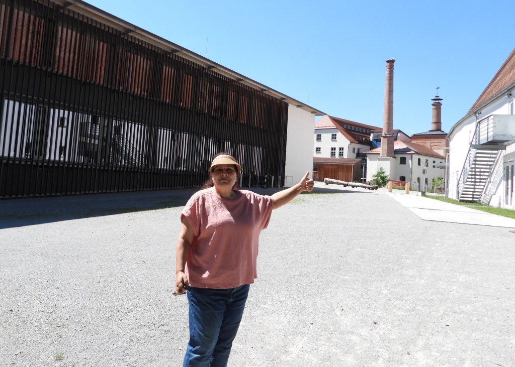 Eine Person steht auf einem kiesigen Weg und zeigt mit dem Daumen nach oben, während im Hintergrund historische Gebäude und Schornsteine zu sehen sind. Der Himmel ist blau und sonnig.