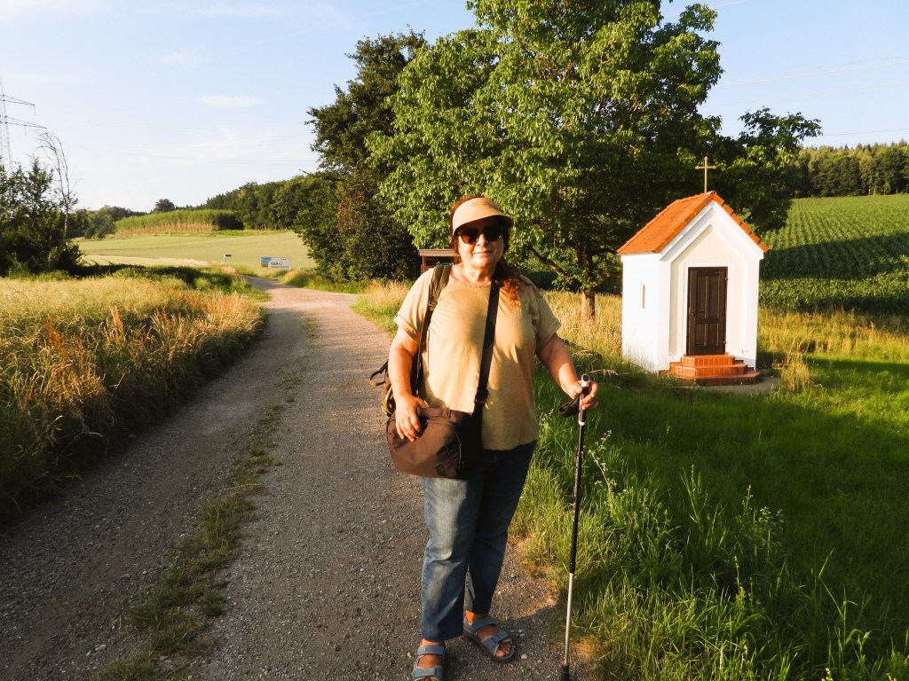 Eine Person mit einer Sonnenbrille und einem Hut steht auf einem Schotterweg, neben einer kleinen Kapelle. Im Hintergrund sind grüne Felder und Bäume zu sehen.