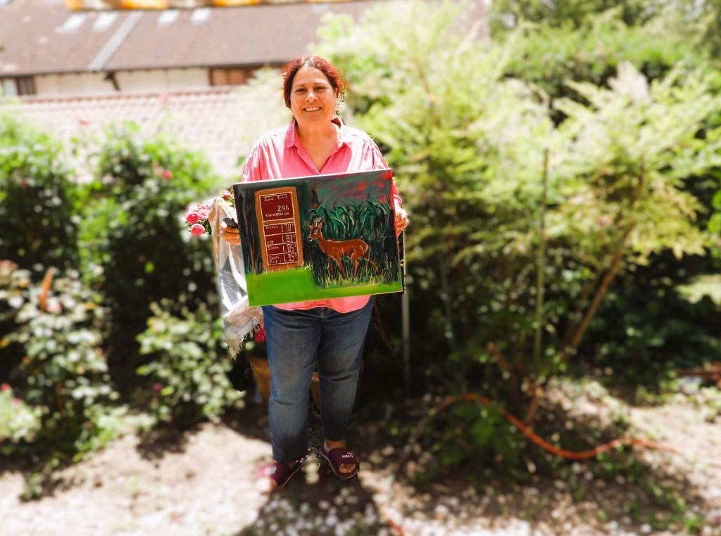 Eine Frau steht in einem Garten und hält ein Gemälde eines Rehs in der Hand. Sie trägt ein pinkes Hemd und lächelt.