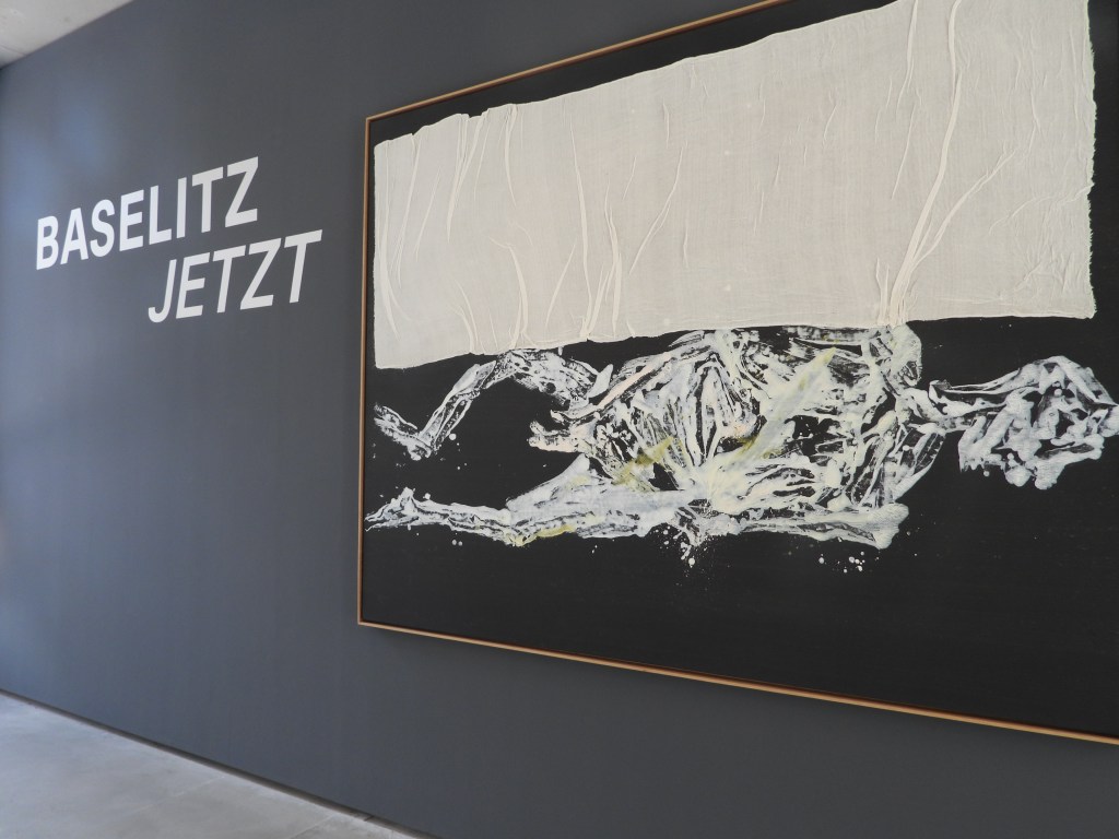 Eine Kunstinstallation mit dem Titel "Baselitz Jetzt" an einer Wand. Das Bild zeigt eine abstrakte Darstellung mit menschlichen Figuren in hellen Farben auf einem dunklen Hintergrund.