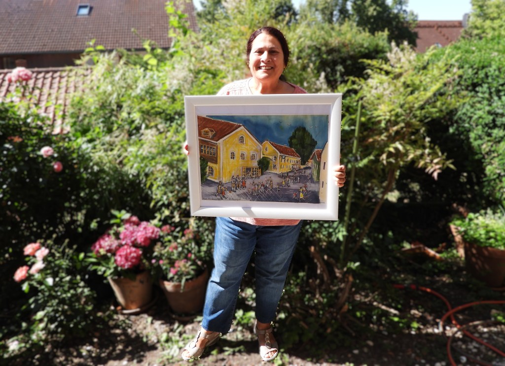 Eine Person hält ein gerahmtes Gemälde mit einer Stadtszene in der Hand, umgeben von einem Garten mit Blumen und grünen Pflanzen.